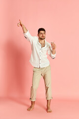 Portrait of handsome young man in stylish clothes posing with cocktail, dancing against pink studio background. Concept of emotions, male fashion, party, fun, celebration, lifestyle. Ad