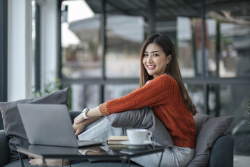 asian young woman listening music with headphone and streaming music from laptop on sofa relaxing at home