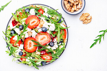 Strawberry and herbs healthy salad with arugula, blueberries, soft white feta cheese and walnuts, white kitchen table, place for text. Fresh useful dish for healthy eating