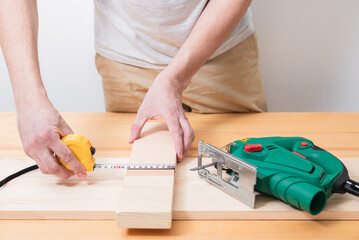 A man works with an electric jigsaw for wood on a wooden table with and without gloves and measures with a tape measure