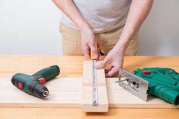 A man works with a jigsaw and a screwdriver on a wooden table with and without gloves, also measures with a tape measure