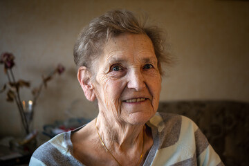 Portrait of an elderly white woman at her home with a sofa ad a flower in the background
