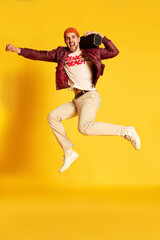Fototapeta na wymiar Portrait of emotive happy young man posing in stylish casual clothes, jumping with music player against yellow studio background. Concept of emotions, male fashion, lifestyle, facial expression. Ad