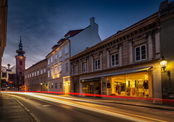 Fototapeta na wymiar View on the main road, historical houses, shops and church tower in the Austrian town of Hainburg during evening with light trails from cars