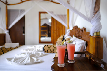 cozy bedroom with mosquito net, white mesh canopy, two glasses of fresh juice by wooden bed in...