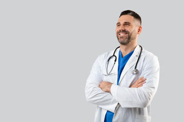 Fototapeta premium Confident male doctor in uniform posing with folded arms over grey studio background, looking at free space