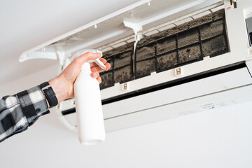 A man's hand sprays a cleaning liquid on the moldy filters of an air conditioner.