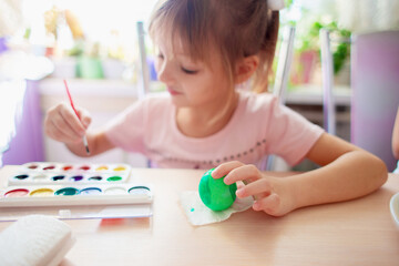A girl sits in the kitchen and paints Easter eggs with watercolor paints A child draws patterns on an Easter egg with a brush