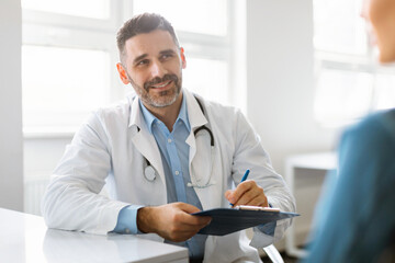 Cheerful middle aged male doctor in white coat with stethoscope consulting patient in office clinic interior - Powered by Adobe