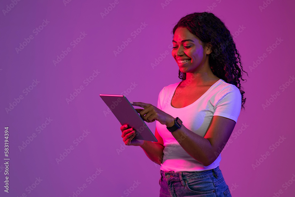 Poster smiling black female using digital tablet while standing in neon light
