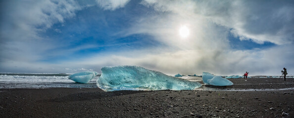 A view of an azure-colored block of ice from an iceberg carried by a storm onto black sand, and in the background, photographers take a photo of a storm on the coast. Jokursarlon, Iceland.