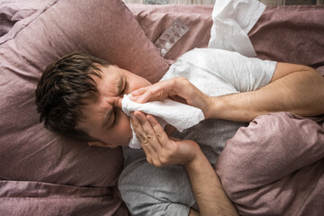 Ill caucasian young man sick with flu, cold or virus blowing running nose, got fever caught cold, sneezing in tissue