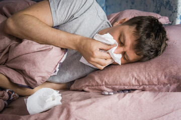 Fototapeta na wymiar Ill caucasian man in bed with virus, flu or cold blowing running nose surrounded by used tissues