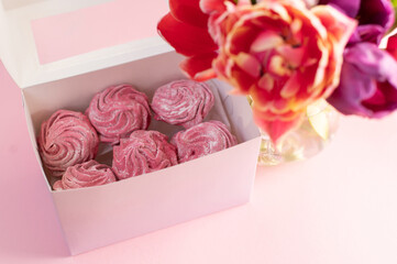 Pink marshmallow lies in a gift box near a bouquet with tulips on a pink background