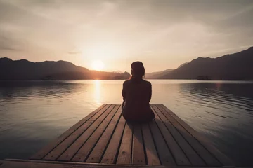 Fotobehang  a person sitting on a dock looking out over a lake at sunset or sunrise or sunset, with mountains in the distance, and a boat in the foreground.  generative ai © Oleg