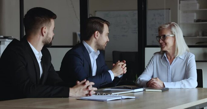 Millennial 35s businessman and elderly 60s businesswoman shake hands finish collaboration at group negotiations gathered together at modern office boardroom. Partnership, teamwork, business etiquette