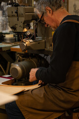 vertical photo with side view of a leather craftsman sitting while sewing a leather back of a chair in his traditional workshop in the historic center of rome, italy