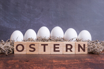 Ostern auf Holzwürfeln vor weißen Eiern, die in Gras auf einem Holztisch liegen.
