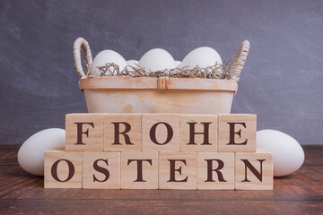 Frohe Ostern auf Holzwürfeln vor einem Holzkorb mit weißen Eiern, auf einem Holztisch.