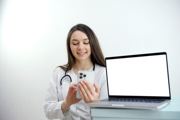 friendly female doctor over white background with a laptop. woman doctor working on the phone answering the call in front of her laptop with white screen space for advertising High quality photo - obrazy, fototapety, plakaty