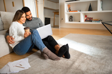 Cheerful international millennial couple looking at laptop, have video call, enjoy coffee cup, sit on floor