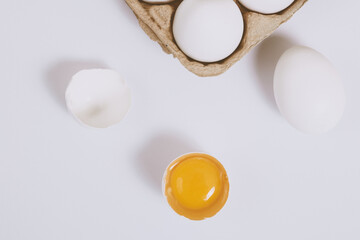 Cracked egg half with a yolk inside near the egg tray with eggs on bright background. Selective focus. Concept scene.  Top view