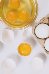 Cracked egg half with a yolk inside near the egg tray with eggs and glass bowl with egg yolks and egg whites on bright background. Close up, selective focus. Top view.