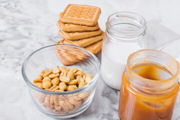 Ingredients for cooking. Stack of cookies, caramel, heap of peanuts, cream in glassware on table. Selective focus.