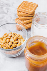 Ingredients for cooking. Stack of cookies, caramel, heap of peanuts, cream in glassware on table. Selective focus.