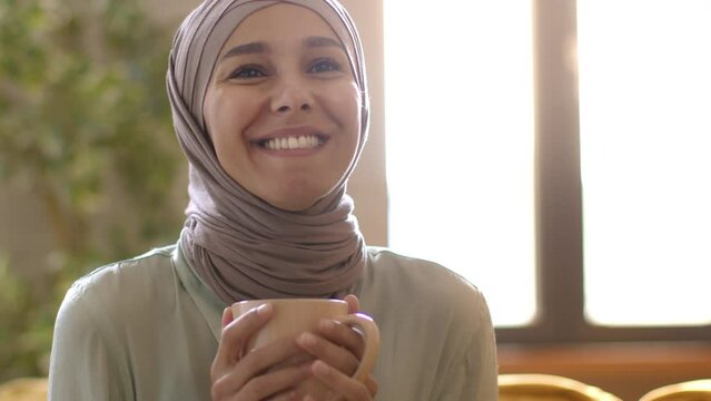 Young positive middle eastern lady in hijab enjoying aromatic fresh hot coffee, tracking shot, empty space