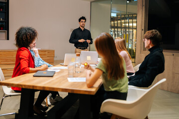 Wide shot of professional male speaker giving corporate presentation for diverse businesspeople at meeting room, presents new business plan. Startup team sitting at boardroom at office listening boss.