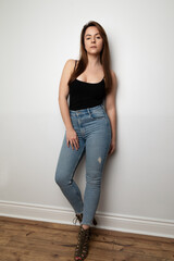 Studio portrait of a beautiful Ukrainian model in her twenties standing agains at white wall. She is wearing high waisted jeans and a black tank top. She has long brown hair and brown eyes. 