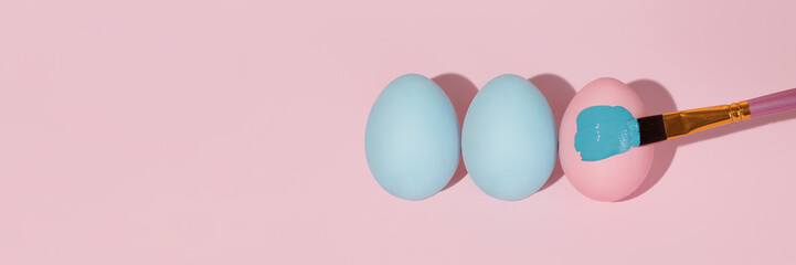 Brush paints a pink Easter egg with blue gouache. Three easter colored eggs lie in a row flat lay....