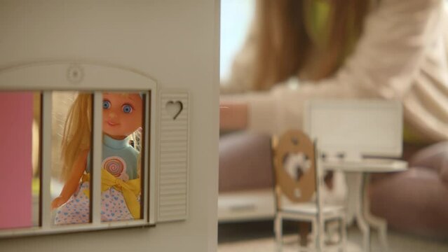 Blond doll standing at window of wooden toy house, unrecognizable teenage girls playing on background, fancy artificial furniture, active imagination, sunny weekend at home, role games. HQ 4k footage