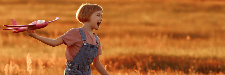 The girl runs across the summer field and launches a toy plane into the sky. A child plays aviator or pilot in a field at sunset, a happy childhood. Banner for website header design with copy space.