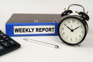 On a white surface, an alarm clock calculator and a folder with the inscription - WEEKLY REPORT