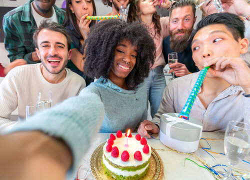 Joyful group of  multiethnic friends taking selfie while having birthday party at home, using mobile phone and drinking champagne