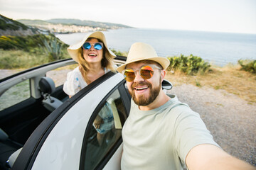 Happy beautiful couple in love taking a selfie portrait driving a convertible car on the road at vacation. Rental cars and vacation