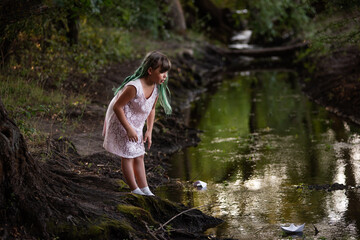 Teenage girl with green hair in dress, stands on the river bank, launches white paper origami boat