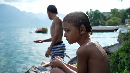 Children by lake drying outside. Pensive kid sitting by water in nature outdoors. Thoughtful child...