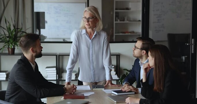 Mature businesswoman, business trainer or mentor leads group meeting with business partners, staff members or subordinates, tells bout collaborative project, share corporate goals and sales results