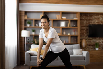 African american woman with dark hair stretching legs during workout at modern living room. Sporty female in activewear smiling and looking at camera while training during free time.
