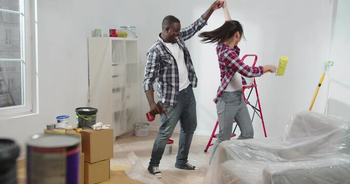 Young cute couple of lovely attractive Asian woman dancing and having fun during renovation of flat with happy joyful African American man. Two beautiful cheerful people renew with fun.