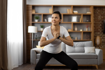 Fototapeta na wymiar Fitness sport woman dressed in active outfit squatting while exercising alone at living room. Multiracial young female training regular at home for staying fit and healthy.
