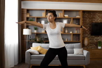 Fototapeta na wymiar Focused american woman in sport clothes standing at cozy living room and practising yoga exercise on mat. Concept of concentration, relaxation and workout.