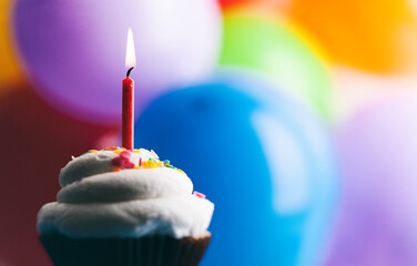 Cupcake With One Birthday Candle Against Colorful Balloon Background