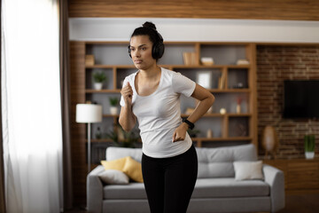 Fototapeta na wymiar Active multiracial woman with dark hair enjoying music in wireless headphones while training at living room. Fitness young lady in sport clothes using modern gadget for workout.