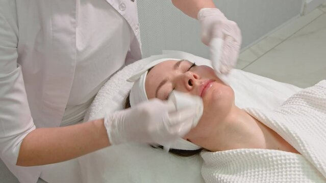 The hands of a beautician cleanse the skin. Facial skin care. A young woman at the cosmetic procedure. Facial cleansing procedure in a cosmetology clinic.