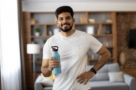 Sporty indian man smiling sincerely on camera while standing in living room with sport bottle of water in hand. Handsome guy wearing active clothes during exercises.