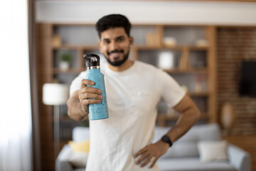 Sporty indian man smiling sincerely on camera while standing in living room with sport bottle of...
